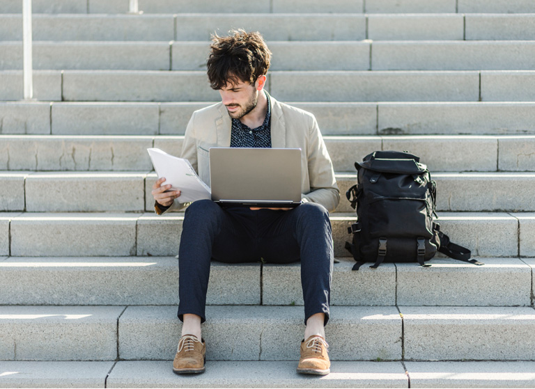 flumoxed student inspecting a piece of paper while using laptop and sitting beside a backpack